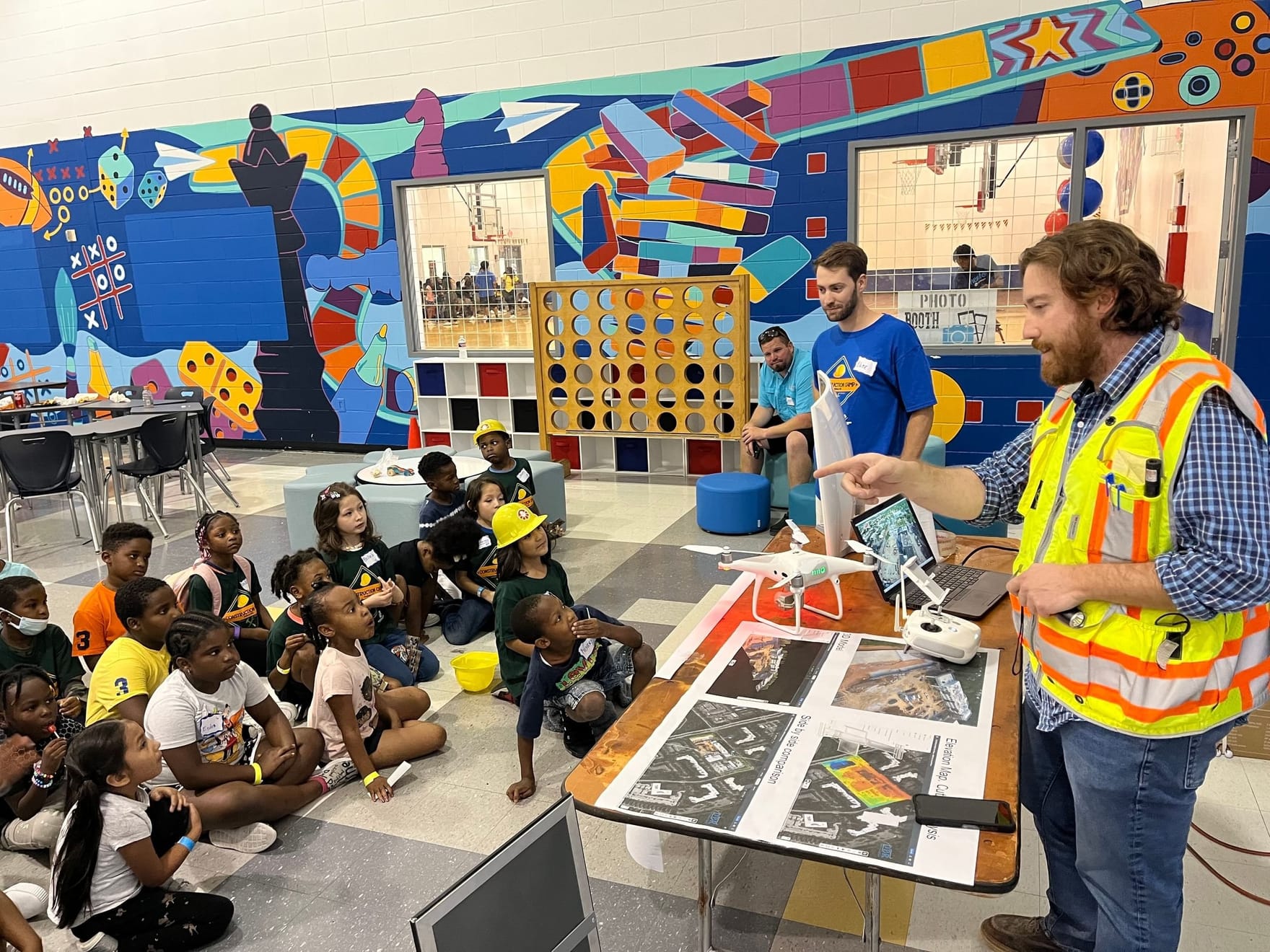 An individual wearing a safety vest stands behind a desk and speaks to a group of children in a classroom.