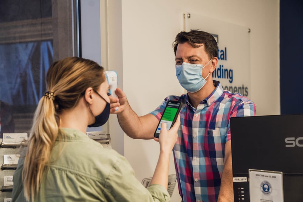 Two people wearing face masks and one taking the temperature of another