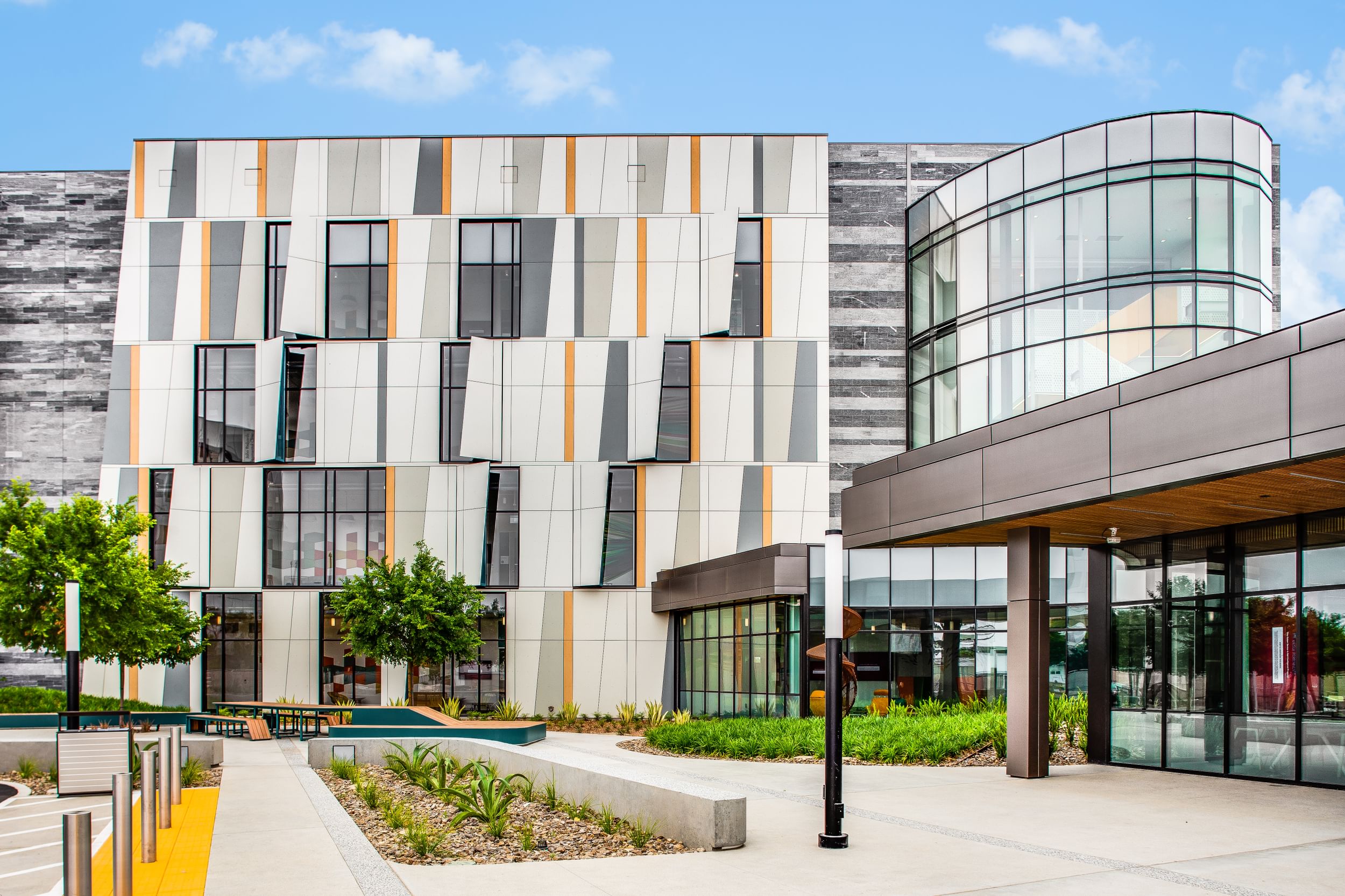 Modern building exterior with a mix of vertical panels in gray, white, and orange tones, large windows, and a curved glass section on the right, surrounded by landscaped greenery.