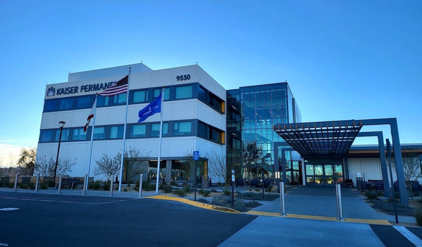 Completed modern building with Kaiser Permanente signage, glass entrance, and three flagpoles displaying the U.S., California, and Kaiser flags. The structure has white exterior panels and a landscaped front area under a bright blue sky