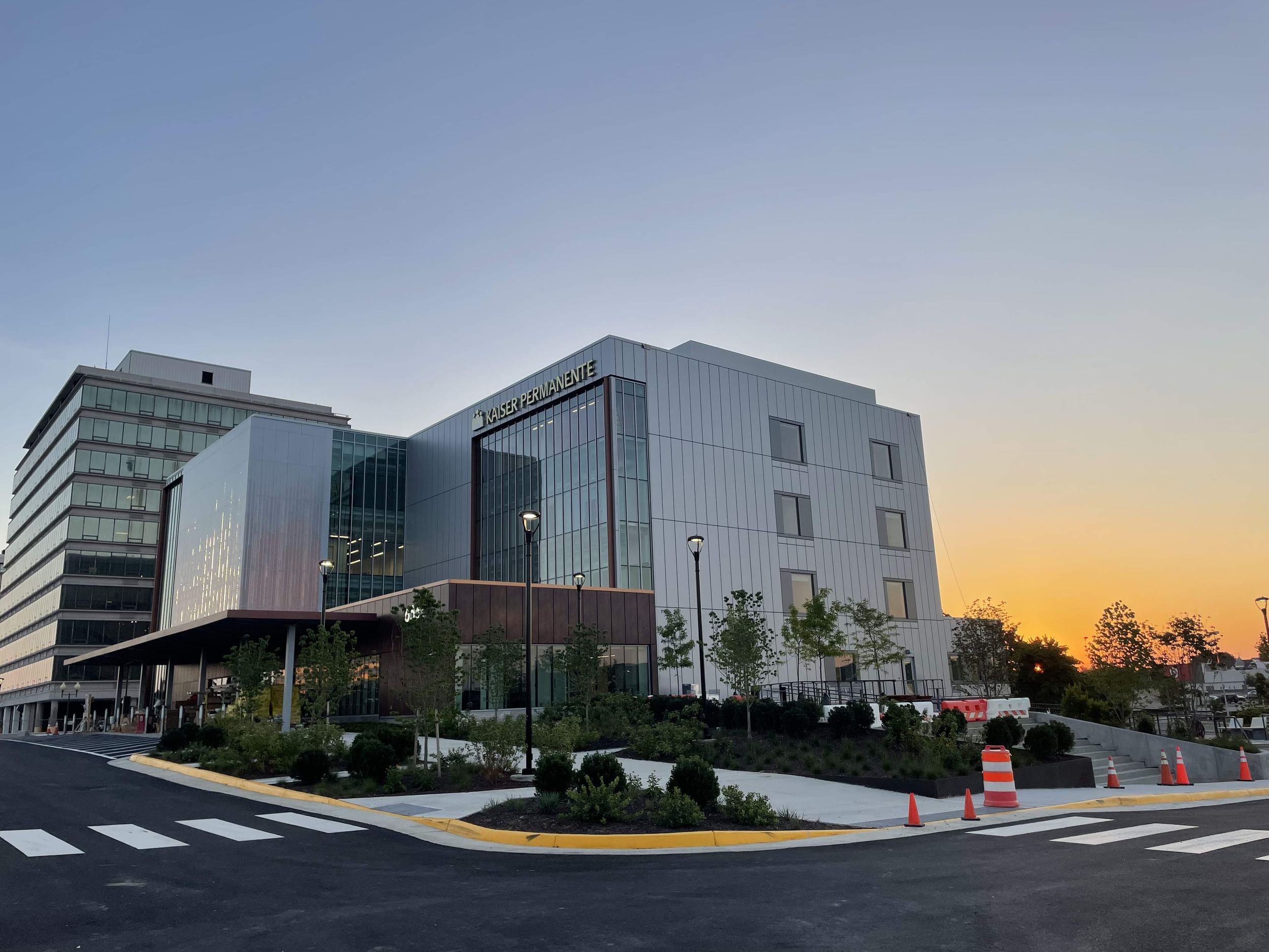 Street view of a finished Kaiser Permanente facility with metal panel exterior, glass sections, and landscaped surroundings under a partly cloudy sky.