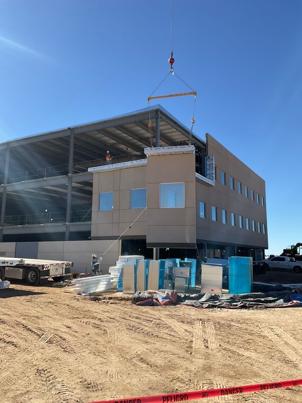 A crane installs a prefabricated exterior panel on a multi‑story building under construction, with materials staged on the ground below.