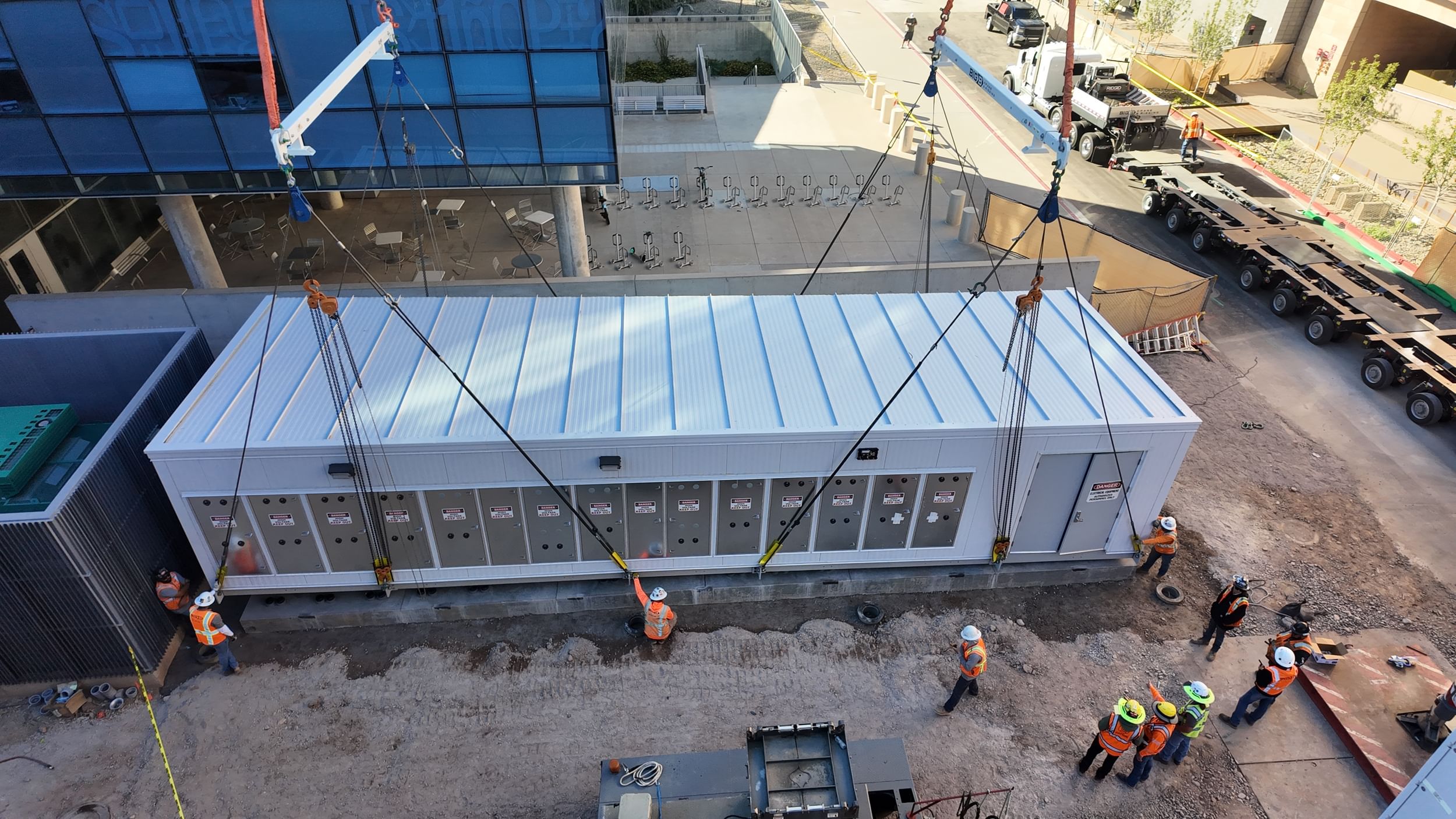 Overhead view of a prefabricated electrical skid being lowered onto its foundation by crane as construction personnel monitor and guide the placement.