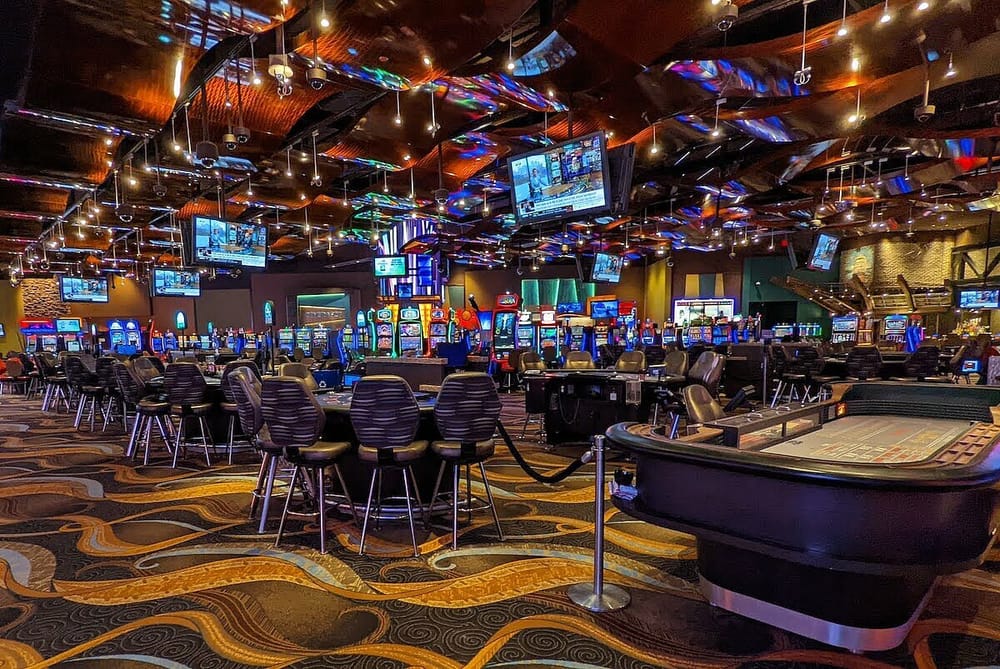 A casino floor with slot machines and poker tables. There are tvs hanging above the tables
