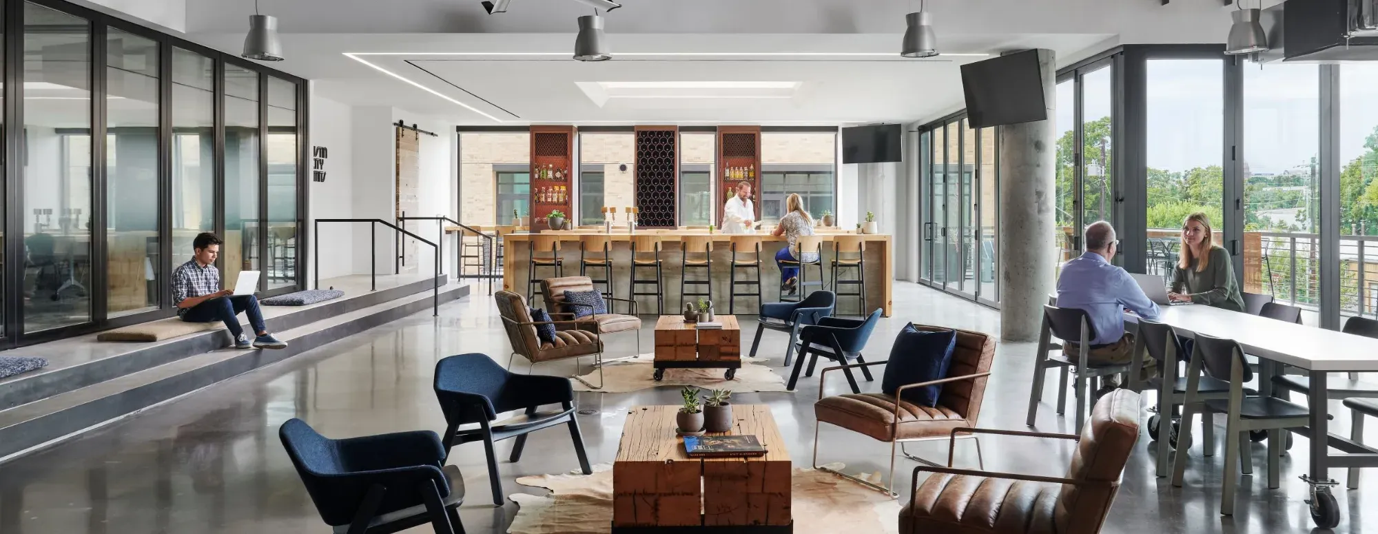 Modern open office lounge with natural light, featuring comfortable seating, a long communal table, and a bar counter. People are working on laptops, sitting in chairs, and having conversations in a collaborative workspace with floor-to-ceiling windows.