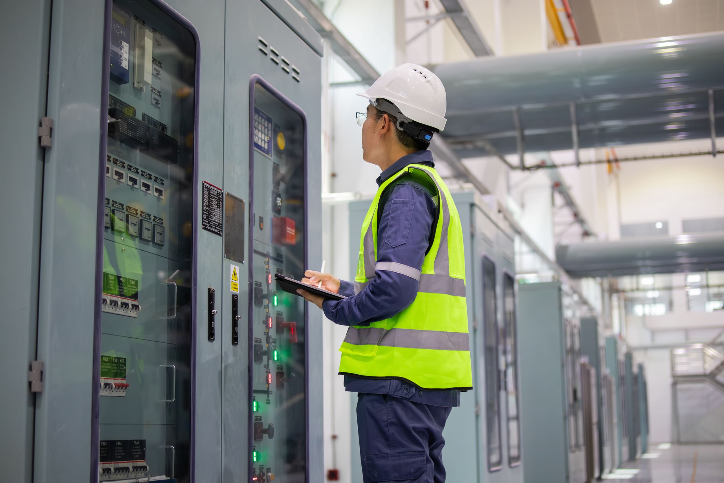Technician inspecting industrial control panels and recording data on a tablet.